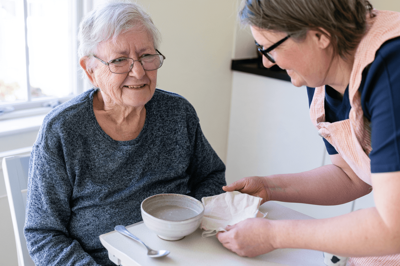 Patient seated upright with a small bowl of soup while a caregiver helps prepare the table.